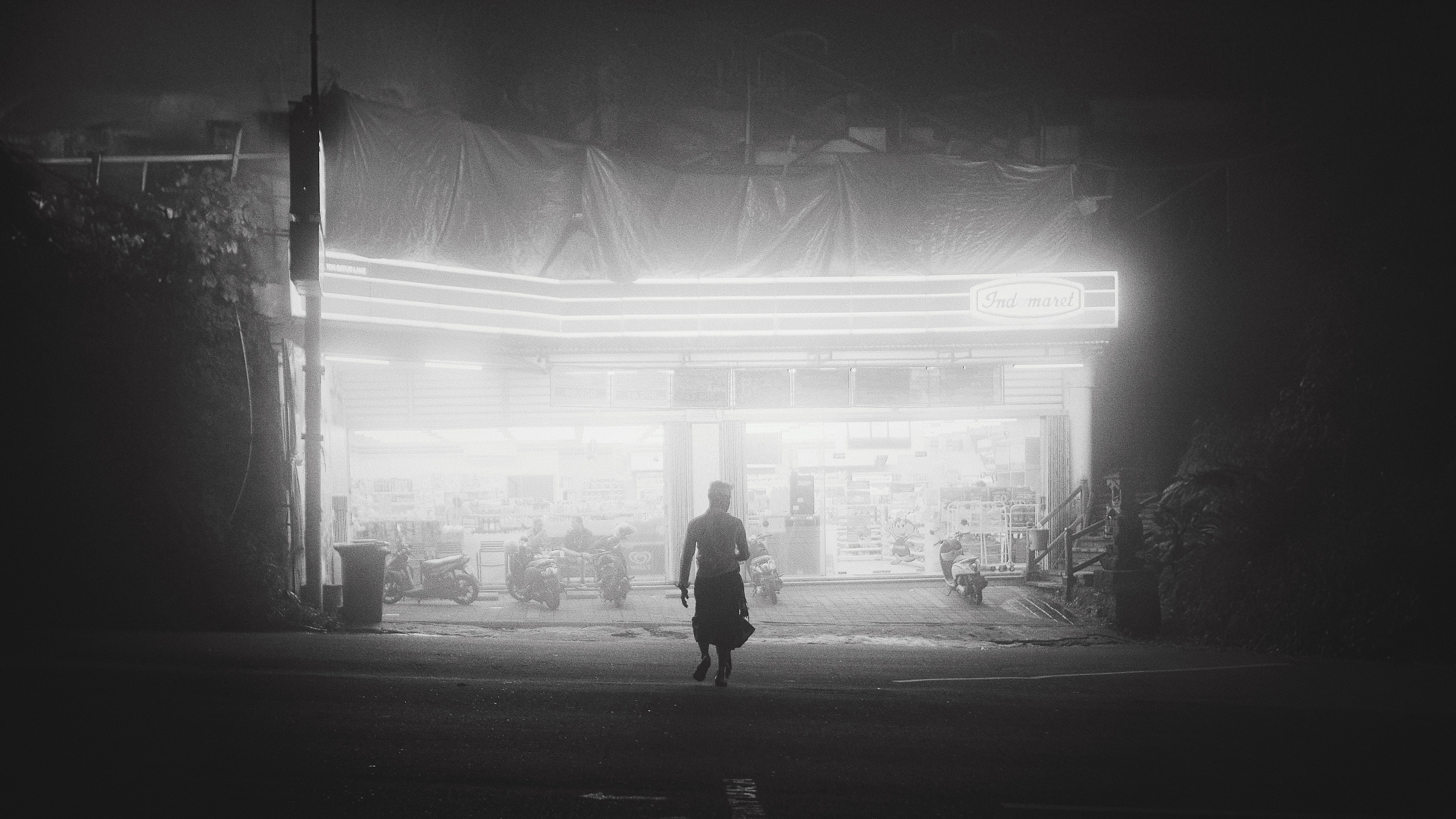 A figure in front of a brightly lit shop at night, in black and white.
