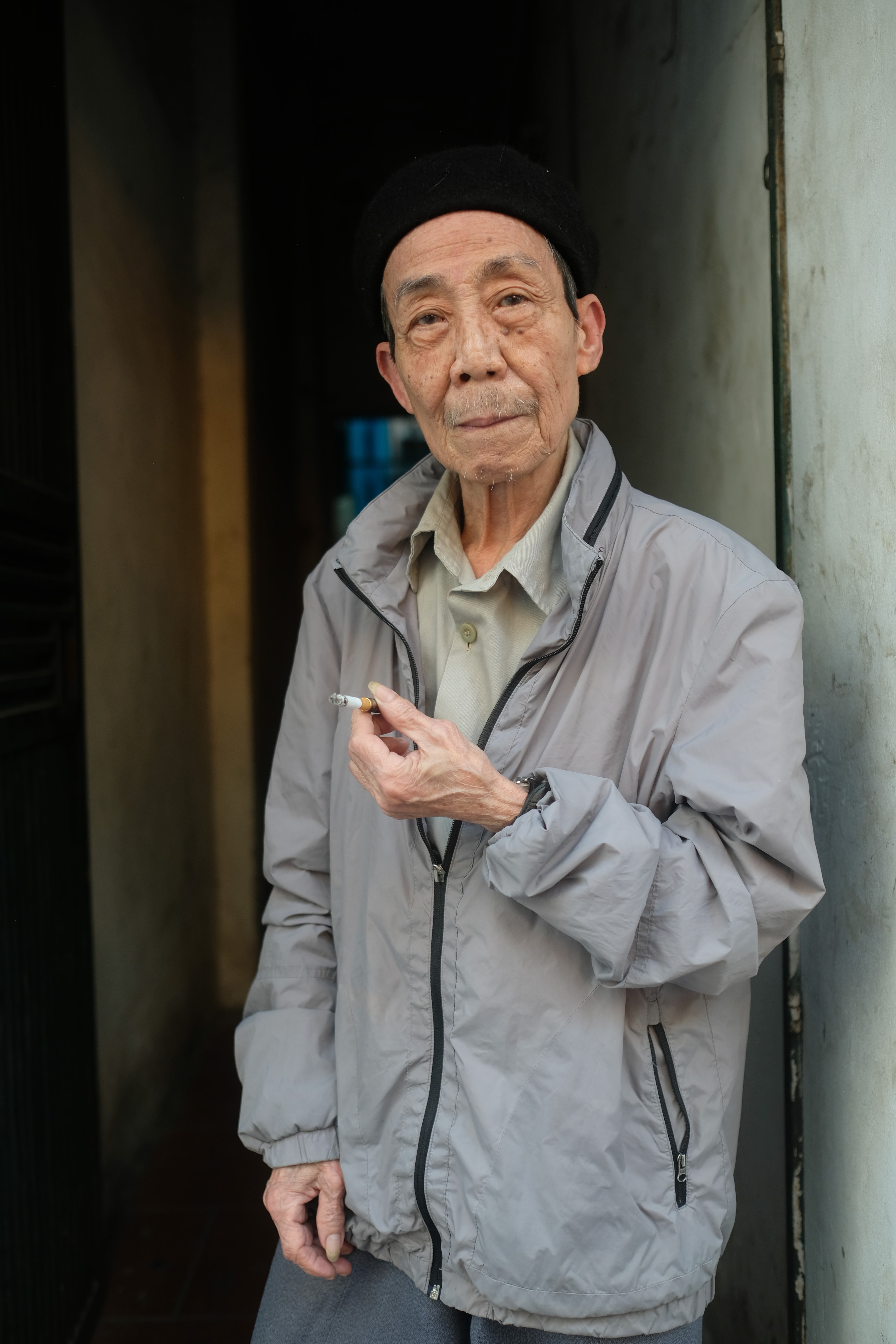 An older man in a grey jacket holding a cigarette in a doorway.