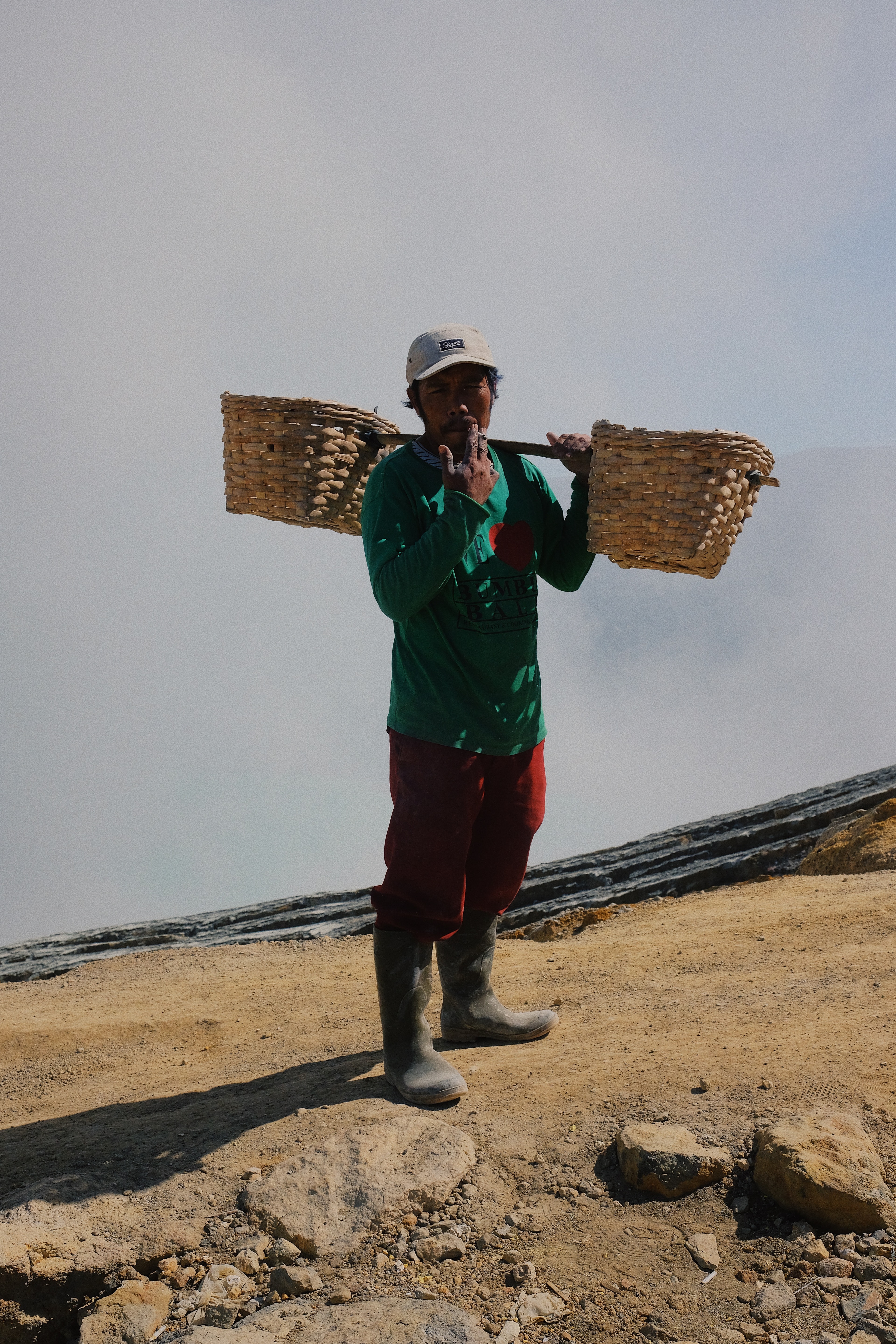 A man carrying two woven baskets on a pole across his shoulders on a rocky slope.