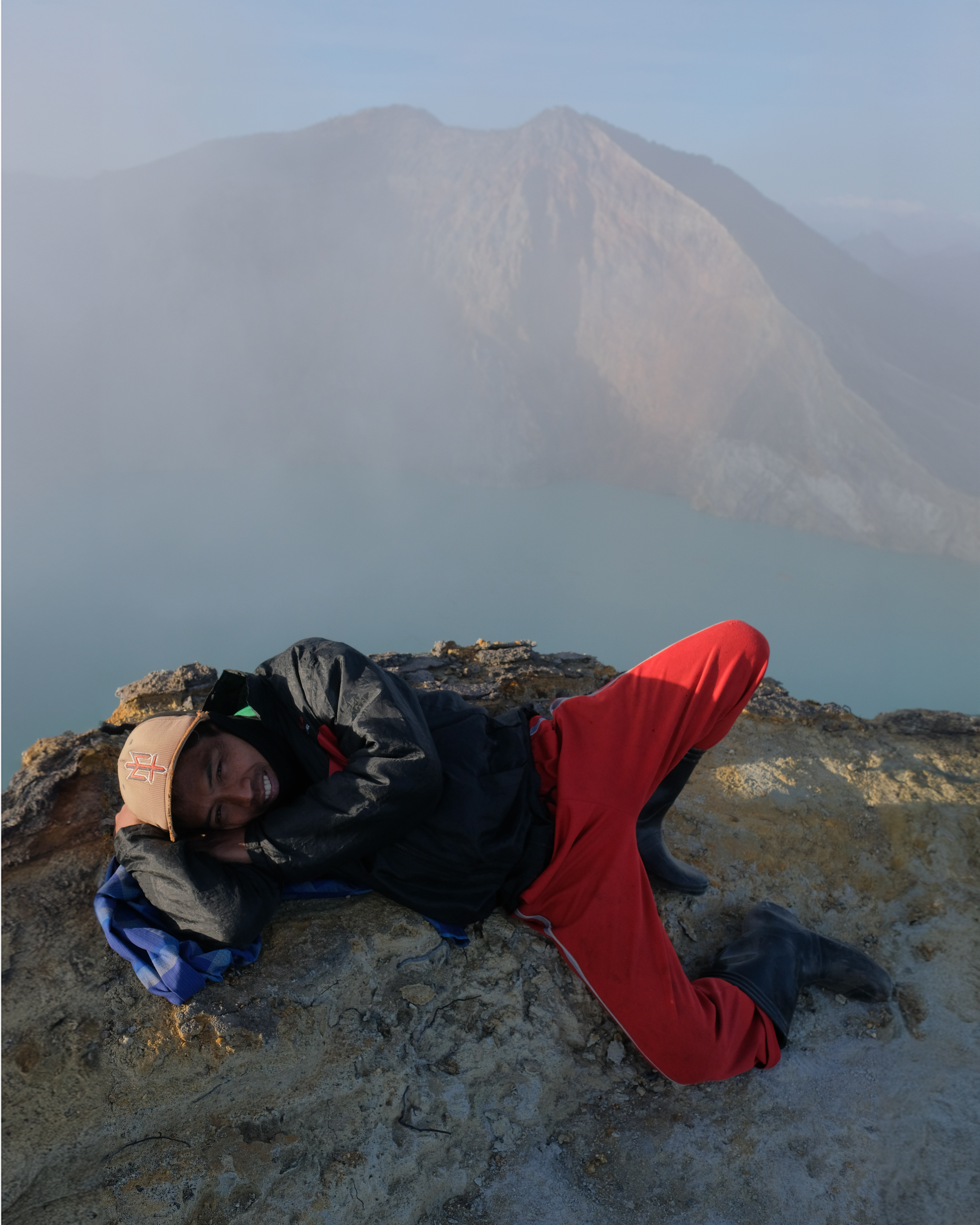 A man in red trousers lying on rocky ground above a crater lake.